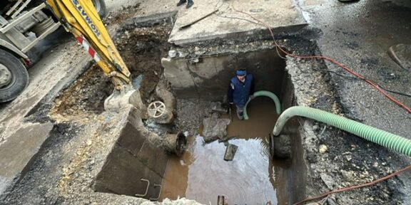 Construction site: a deep excavation with water in the pit, a worker standing inside wearing a blue hard hat/vest, and large hoses looping in and out of the trench.