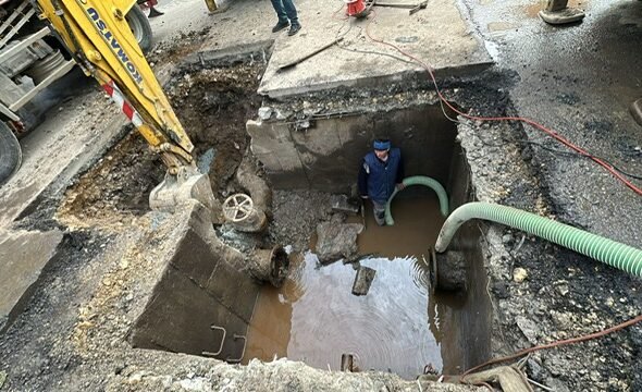 Construction site: a deep excavation with water in the pit, a worker standing inside wearing a blue hard hat/vest, and large hoses looping in and out of the trench.