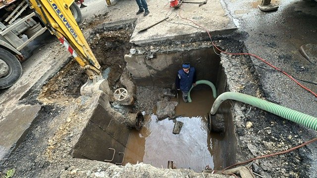 Construction site: a deep excavation with water in the pit, a worker standing inside wearing a blue hard hat/vest, and large hoses looping in and out of the trench.