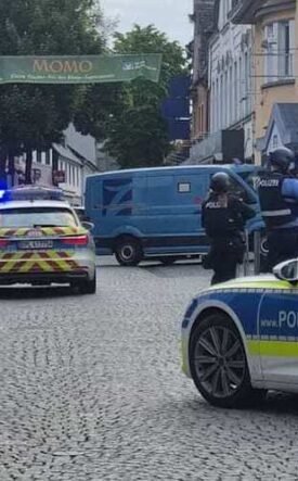 German police cars with blue lights block a cobblestone street; two officers in protective vests stand near a blue van amid shops and pedestrians.