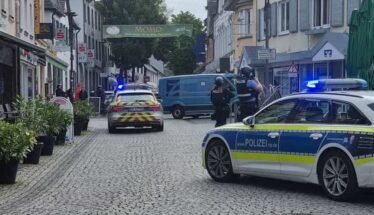 German police cars with blue lights block a cobblestone street; two officers in protective vests stand near a blue van amid shops and pedestrians.