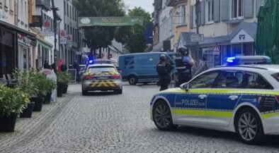 German police cars with blue lights block a cobblestone street; two officers in protective vests stand near a blue van amid shops and pedestrians.