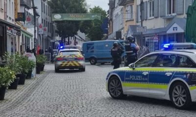 German police cars with blue lights block a cobblestone street; two officers in protective vests stand near a blue van amid shops and pedestrians.