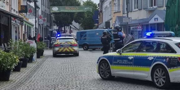 German police cars with blue lights block a cobblestone street; two officers in protective vests stand near a blue van amid shops and pedestrians.