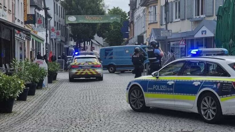 German police cars with blue lights block a cobblestone street; two officers in protective vests stand near a blue van amid shops and pedestrians.