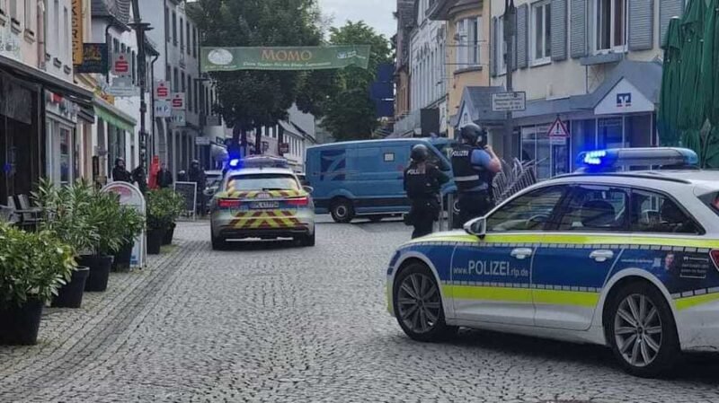German police cars with blue lights block a cobblestone street; two officers in protective vests stand near a blue van amid shops and pedestrians.