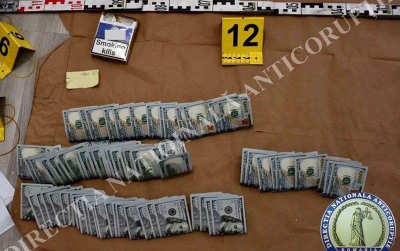 Evidence board showing several bundles of U.S. hundred-dollar bills laid out in rows, with a 'Smoking kills' cigarette pack and a yellow 12 marker nearby.