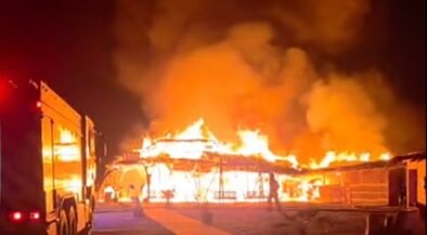 Building is engulfed in flames at night with fire trucks nearby as emergency responders tackle a large blaze.