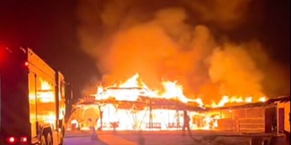 Building is engulfed in flames at night with fire trucks nearby as emergency responders tackle a large blaze.