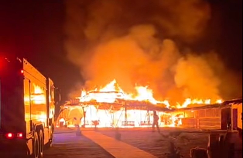 Building is engulfed in flames at night with fire trucks nearby as emergency responders tackle a large blaze.