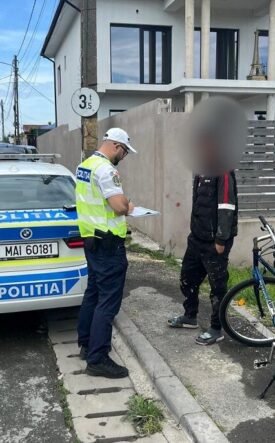 Policeman in a reflective vest writes on a clipboard beside a police car on a residential street.