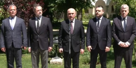 Five men in dark suits stand outdoors around a microphone, appearing to deliver a formal address.