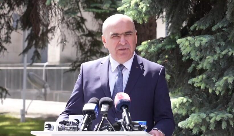 Man in a navy suit delivering a press conference outdoors, with multiple microphones on a podium in front of him.