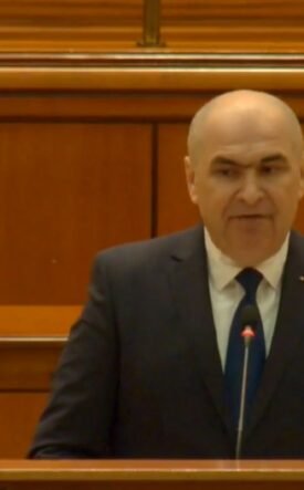 Bald man in a dark suit delivering a speech from a wooden podium in a formal chamber.