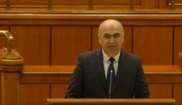 Bald man in a dark suit delivering a speech from a wooden podium in a formal chamber.