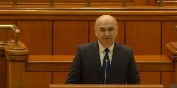 Bald man in a dark suit delivering a speech from a wooden podium in a formal chamber.