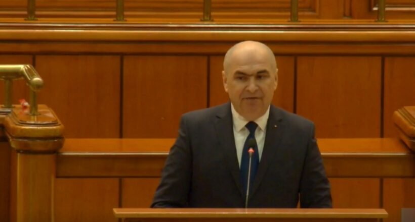 Bald man in a dark suit delivering a speech from a wooden podium in a formal chamber.
