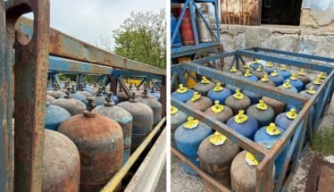 Rows of blue and rusty gas cylinders with yellow valve caps stored in a weathered metal rack outdoors.