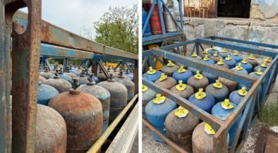Rows of blue and rusty gas cylinders with yellow valve caps stored in a weathered metal rack outdoors.