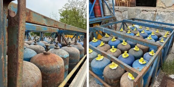 Rows of blue and rusty gas cylinders with yellow valve caps stored in a weathered metal rack outdoors.