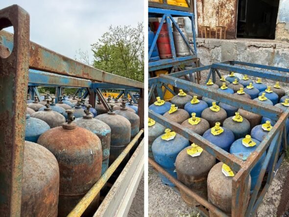 Rows of blue and rusty gas cylinders with yellow valve caps stored in a weathered metal rack outdoors.