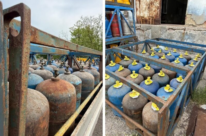 Rows of blue and rusty gas cylinders with yellow valve caps stored in a weathered metal rack outdoors.