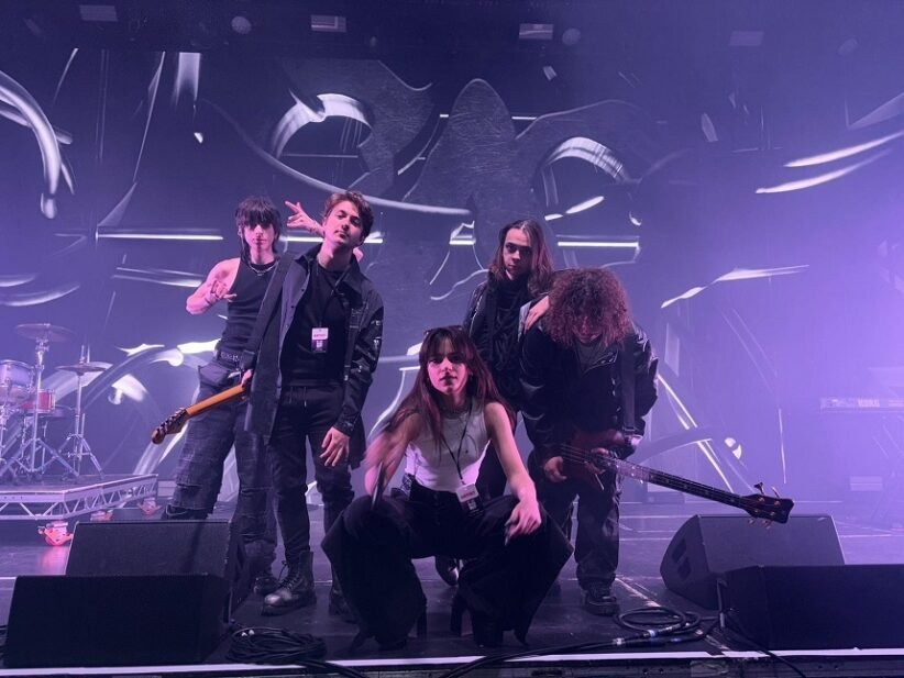 Five-member rock band poses on a stage bathed in purple lighting, with guitars and drums visible in the background.