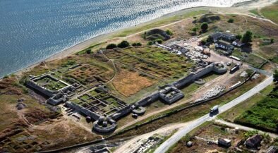Aerial view of an old fortress with stone walls along a coastline, showing circular towers and grassy ruins near a road and small buildings nearby.
