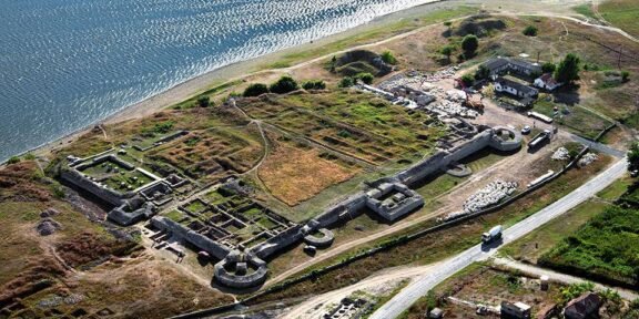 Aerial view of an old fortress with stone walls along a coastline, showing circular towers and grassy ruins near a road and small buildings nearby.