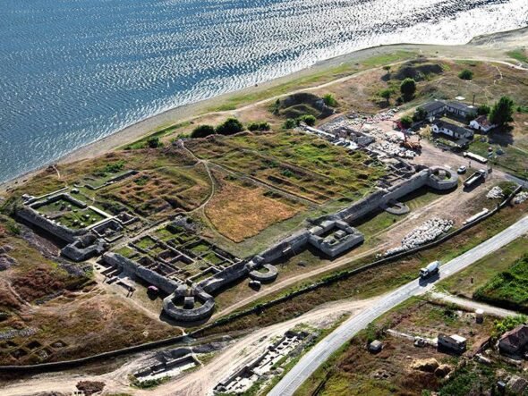 Aerial view of an old fortress with stone walls along a coastline, showing circular towers and grassy ruins near a road and small buildings nearby.
