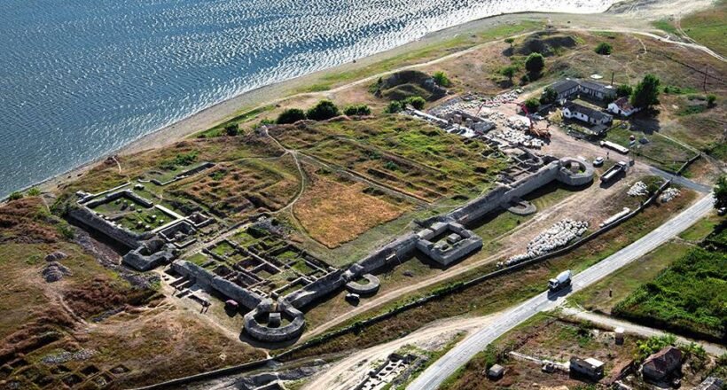 Aerial view of an old fortress with stone walls along a coastline, showing circular towers and grassy ruins near a road and small buildings nearby.