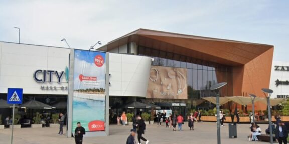 People walk and gather outside a modern shopping mall with a white façade and a copper-colored angular entrance. retailers and banners are visible outside the entrance, City Mall.