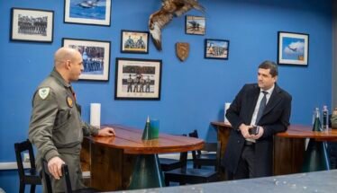 Two men talk at a wooden table in a blue-walled room with framed aviation photos and a mounted hawk between them.