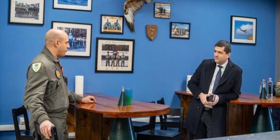 Two men talk at a wooden table in a blue-walled room with framed aviation photos and a mounted hawk between them.