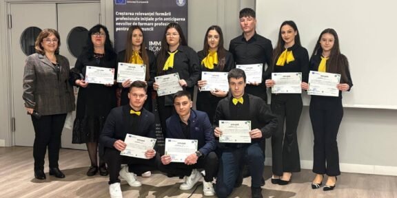 Group of students and two adults posing with certificates at a formal ceremony; some students wear yellow scarves.