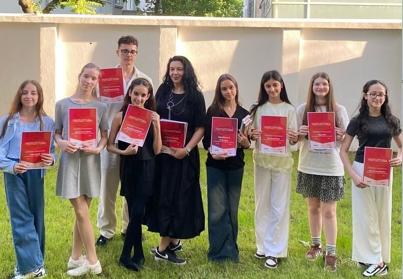 Eleven students and a teacher pose on a lawn, each holding a red certificate, celebrating a ceremony outdoors.
