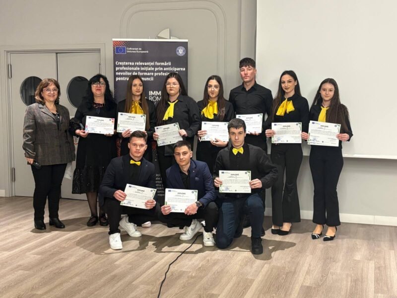 Group of students and two adults posing with certificates at a formal ceremony; some students wear yellow scarves.