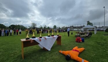 Outdoor awards ceremony: trophies on a table with a white cloth, people lined up in the background, and a person in an orange safety suit lying on the grass in the foreground.