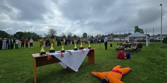 Outdoor awards ceremony: trophies on a table with a white cloth, people lined up in the background, and a person in an orange safety suit lying on the grass in the foreground.