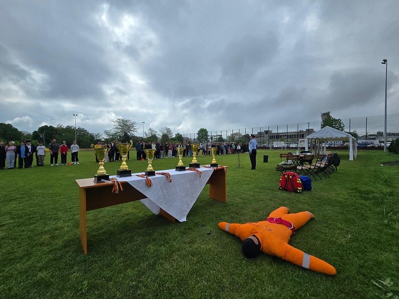 Outdoor awards ceremony: trophies on a table with a white cloth, people lined up in the background, and a person in an orange safety suit lying on the grass in the foreground.