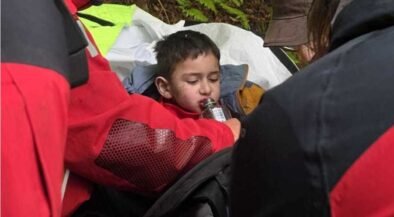 Young boy resting in a makeshift shelter as rescuers in red jackets help him drink from a bottle on a forested trail