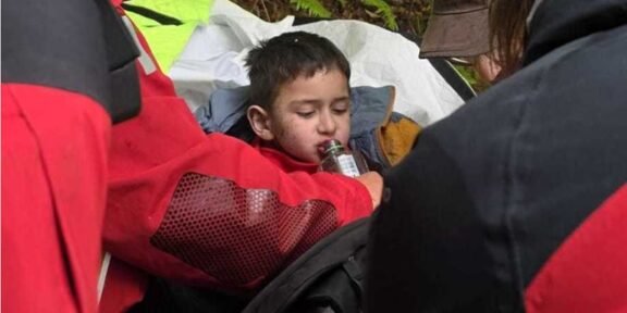 Young boy resting in a makeshift shelter as rescuers in red jackets help him drink from a bottle on a forested trail