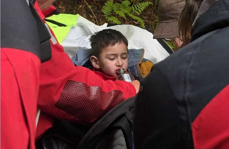 Young boy resting in a makeshift shelter as rescuers in red jackets help him drink from a bottle on a forested trail