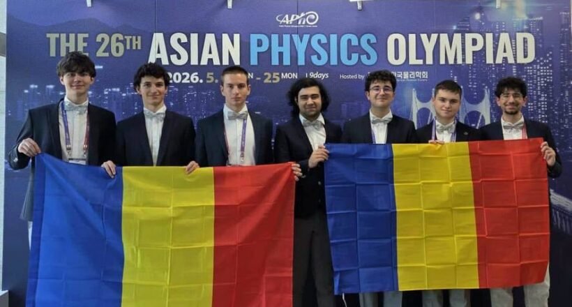 Seven young men in suits pose in front of a blue banner that reads 'The 26th Asian Physics Olympiad,' holding two Romanian flags with blue-yellow-red vertical stripes.