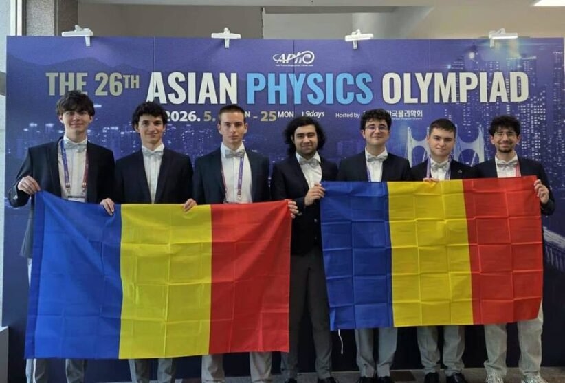 Seven young men in suits pose in front of a blue banner that reads 'The 26th Asian Physics Olympiad,' holding two Romanian flags with blue-yellow-red vertical stripes.