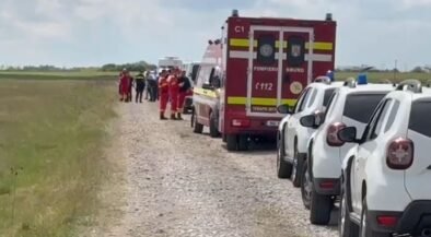 Emergency scene on a rural gravel road: a red ambulance, white parked cars, and firefighters in orange uniforms