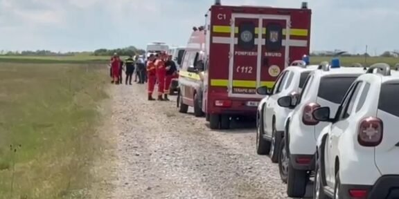 Emergency scene on a rural gravel road: a red ambulance, white parked cars, and firefighters in orange uniforms