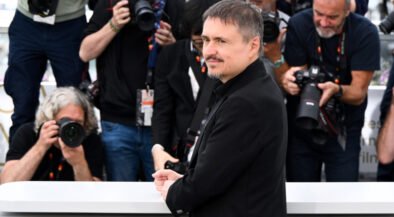 Man in a black coat posing behind a white barrier while photographers surround him.