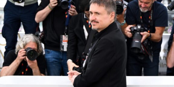 Man in a black coat posing behind a white barrier while photographers surround him.