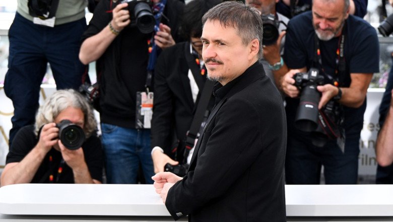 Man in a black coat posing behind a white barrier while photographers surround him.